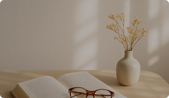 A soft, lifestyle scene showing a coffee table with an open book, reading glasses, a small vase with dried flowers, and gentle morning sunlight casting shadows. The background is airy and uncluttered, perfect for placing text or a CTA